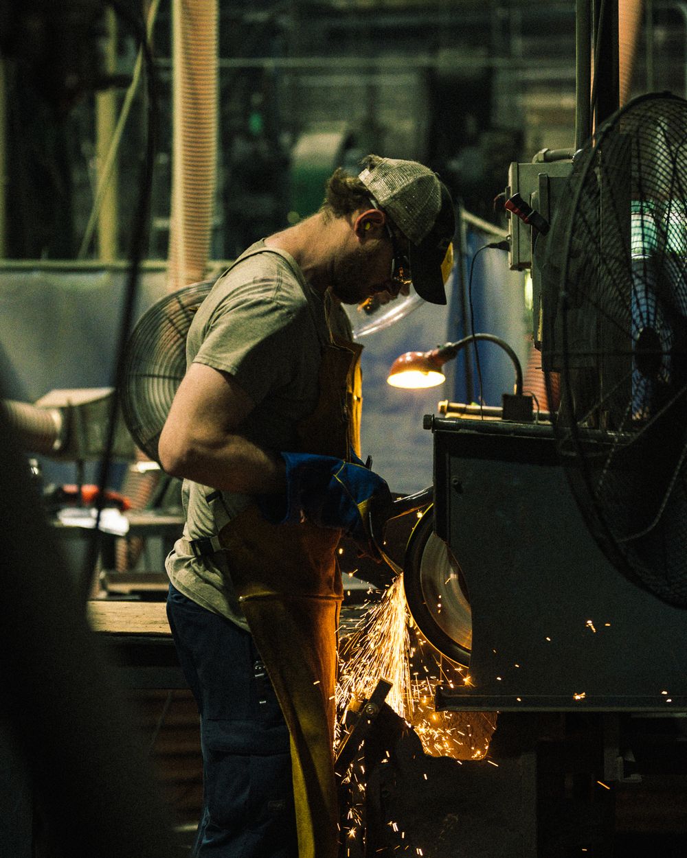A machinist in a leather apron grinds a part as sparks fall off the wheel